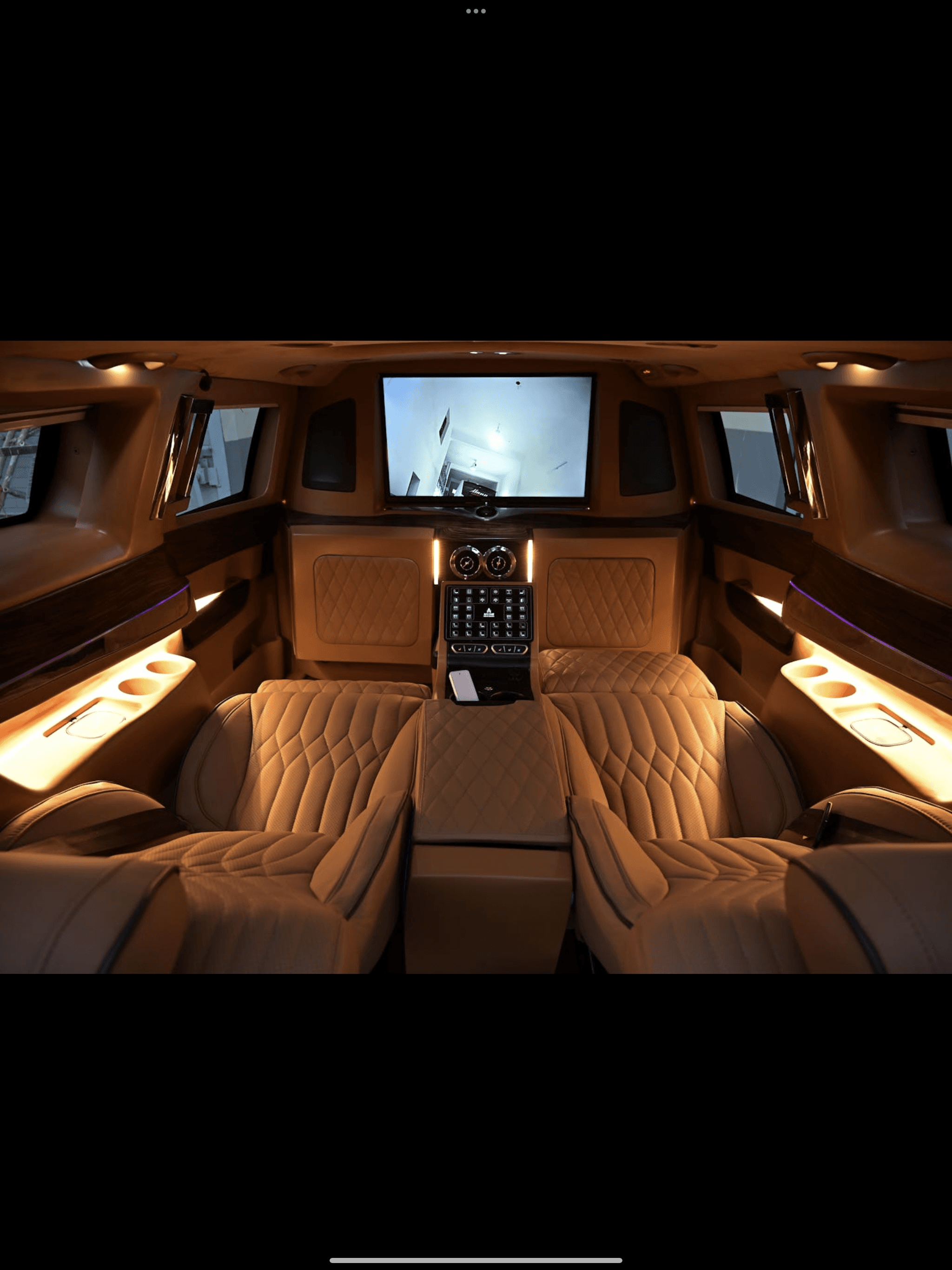Luxurious van interior featuring quilted tan leather seats, a large TV, and warm ambient lighting.